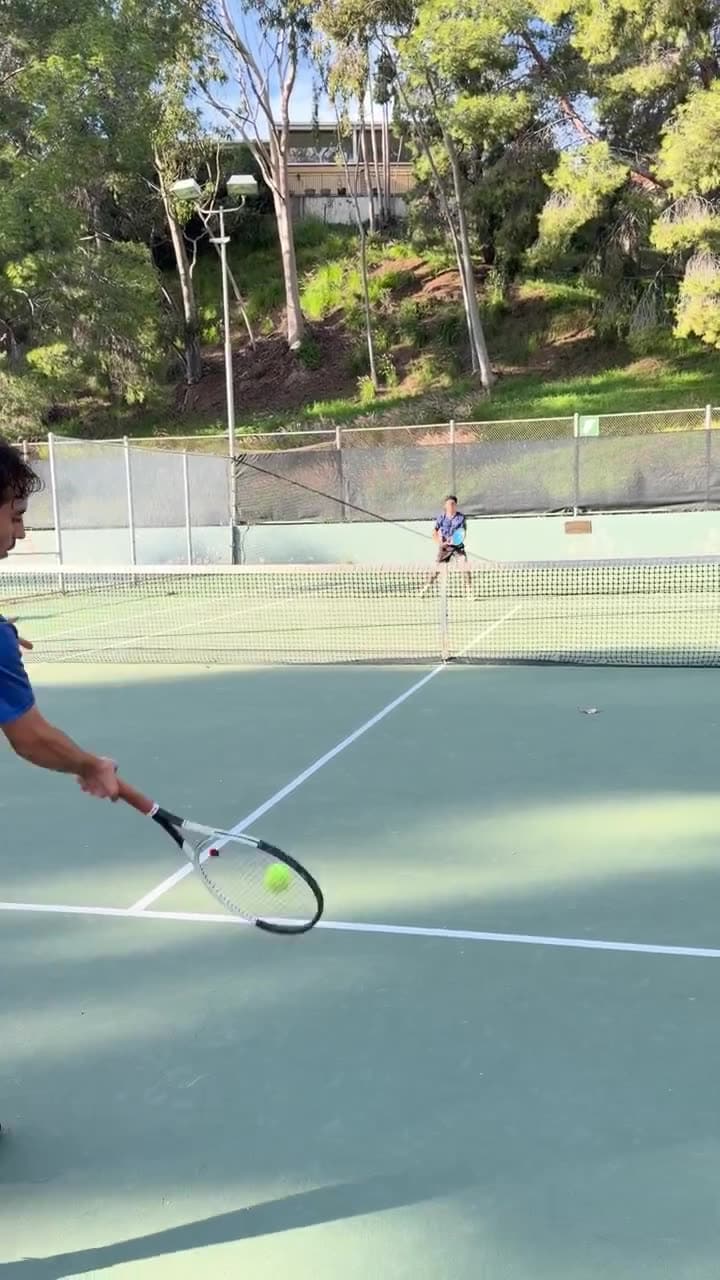 Tennis coach rallying with a player across the net during a lesson on a Los Angeles court